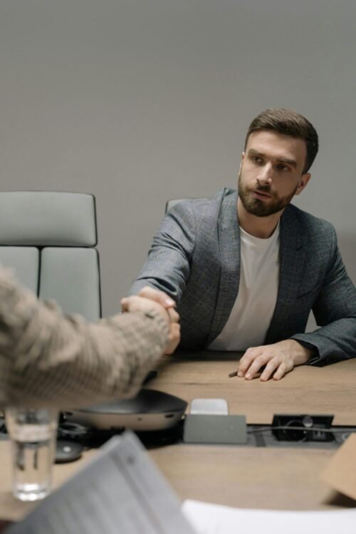 Home Two businessmen in a modern office shaking hands across a table. Professional setting.
